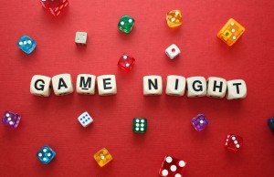 Close up photo with word dice showing the phrase 'Game Night', a slection of colorful dice lay around it on a warm red cardboard backing.
